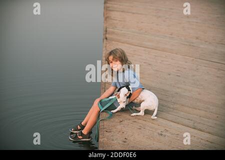 Blonde Junge mit einem Hund an einem See sitzen. Kühler, sonniger Tag. Boy umarmt einen Hund. Unbeschwerte Kindheit. Schutz von Haustiertieren. Stockfoto