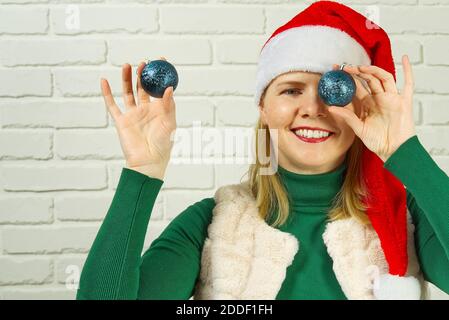 Weihnachtsspaß. Frau in santa roten Hut hält Weihnachtskugeln vor ihren Augen und lächelnd weisen weiße Ziegelwand Stockfoto