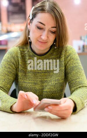 Schöne junge blonde Frau nutzt ihr Handy, während an einem Tisch in einem Restaurant sitzen. Vertikales Foto Stockfoto