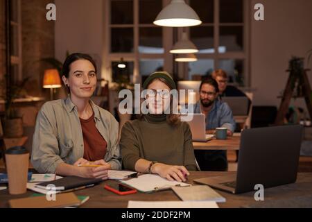 Porträt junger Designerinnen, die während der Arbeit die Kamera betrachten Am Tisch mit Dokumenten und Laptop Stockfoto