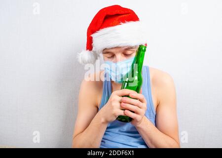 Junger Mann in Weihnachtsmütze und Grippe-Maske in Alkohol Sucht an der Wand im Raum Stockfoto