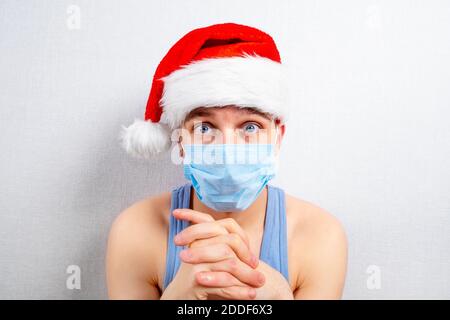 Ängstlicher junger Mann in Weihnachtsmütze in der Grippe-Maske Auf dem Hintergrund der weißen Wand Stockfoto