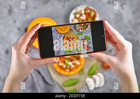 Aufnahme von Lebensmitteln mit Händen halten Handy mit Bild von vegan gebacken Red kuri Squash Gemüse gefüllt mit Paprika, Tomaten und Mushr Stockfoto