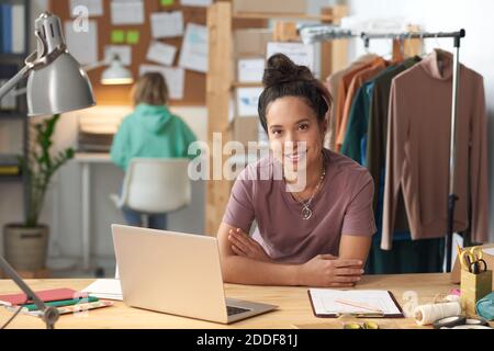 Portrait des Modedesigners, der beim Sitzen die Kamera anlächelt Der Tisch vor dem Laptop in der Werkstatt Stockfoto