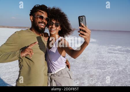 Bild eines glücklichen afroamerikanischen Paares, das sich umarmt und Selfie macht Auf dem Handy im Salztal Stockfoto