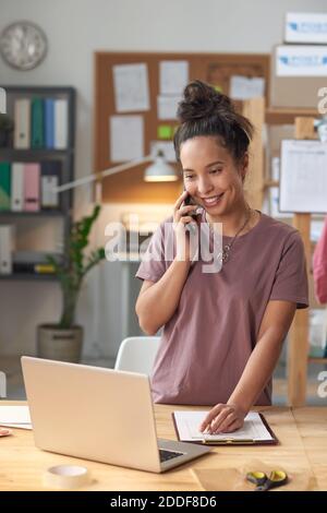 Junge lächelnde Geschäftsfrau hat ein Gespräch auf Handy suchen Am Laptop, der eine Bestellung im Büro annimmt Stockfoto