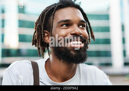 Fröhlicher afroamerikanischer Kerl mit Dreadlocks posiert und lächelt an City Street Stockfoto