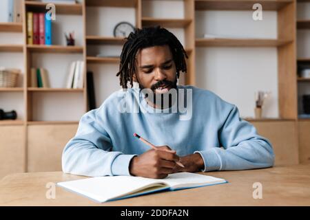 Junger afro amerikanischer Mann, der mit Lehrbuch am Schreibtisch sitzt In der Bibliothek Stockfoto