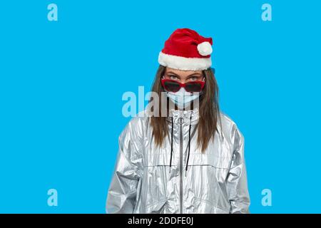 Ernsthafte Frau trägt Weihnachtsmütze und Schutzmaske und rote Sonnenbrille auf blauem Hintergrund im Studio stehen und auf die Kamera schauen. Lady schaut über t Stockfoto