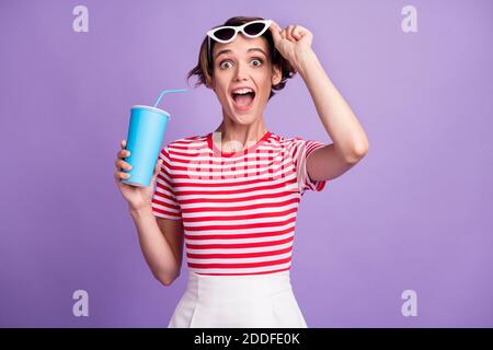 Foto von Dame nehmen Specs halten Soda Tasse offen Mund tragen Sonnenschutz gestreiften T-Shirt isoliert violetten Hintergrund Stockfoto