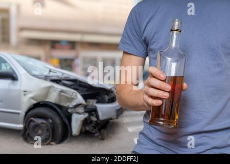 Verkehrsunfall und Alkohol-Konzept. Betrunkener Fahrer hält Flasche mit Alkohol in der Hand. Abgestürztes Auto im Hintergrund. Stockfoto