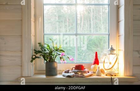 Weihnachtslaterne, Engel, Weihnachtsgnome, Weihnachtskaktus und roter Becher am Fenster eines Holzhauses mit Blick auf den Wintergarten. Stockfoto