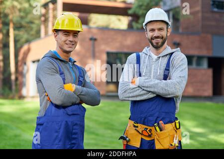 Portrait von zwei hübschen jungen Ingenieuren in Harthüten lächelnd an der Kamera, posiert im Freien mit gekreuzten Armen während der Arbeit an Hütte Bau. Aufbau, Teamarbeit, Partnerschaftskonzept Stockfoto