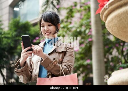 Modische junge asiatische Frau mit Einkaufstaschen Blick auf Handy Lächelnd Stockfoto