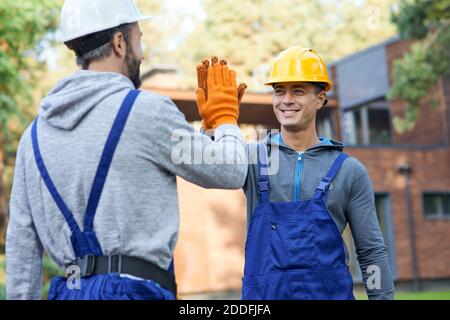 Zwei positive junge Ingenieure in Hartmützen lächeln einander an, geben hoch fünf während der Arbeit auf Hütte Baustelle im Freien. Aufbau, Teamarbeit, Partnerschaftskonzept Stockfoto