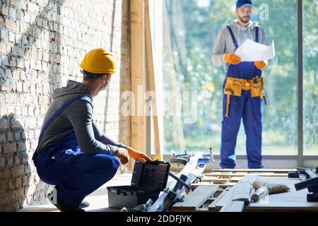 Junger Ingenieur mit Overalls und Hartmütze, der auf der Baustelle des Landhauses arbeitet, Picking Instrument aus der Werkzeugkiste mit verschwommenem Kollegen im Hintergrund an einem sonnigen Tag. Gebäudekonzept Stockfoto