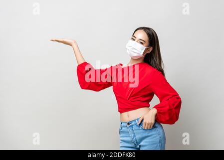 Asiatische Frau trägt medizinische Maske und offene Hand zu leeren Platz beiseite auf hellgrauem Studiohintergrund Stockfoto