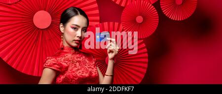 Portrait der schönen asiatischen Frau trägt traditionelle Cheongsam Qipao Kleid Zeigt Kreditkarte in der Hand auf rotem orientalischem Banner Hintergrund Für Chinesisch Stockfoto