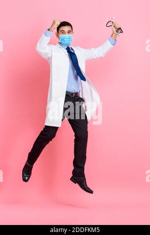 Asiatische männliche Arzt trägt medizinische Maske Heben Fäuste und springen Isoliert in rosa Studio Hintergrund