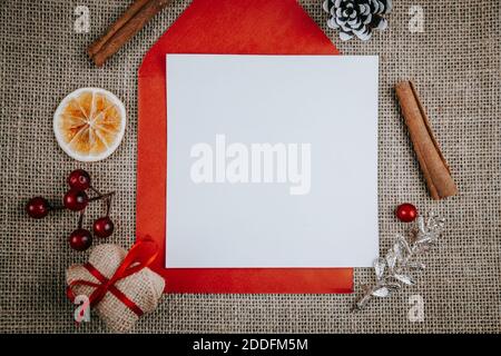 Mockup unbeschriftete Grußkarte, roter Umschlag, Geschenkbox, Zimtstangen. Draufsicht flach Lay gestylt Weihnachten mockup auf einem rustikalen rauen Leinen Hintergrund. Stockfoto