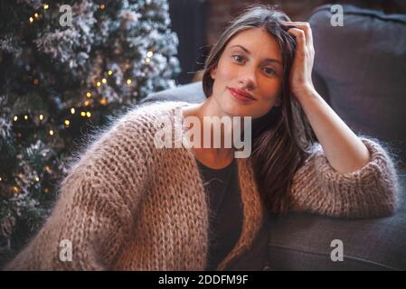 Porträt einer jungen schönen Brünette, die im Wohnzimmer bei einem Weihnachtsbaum in einem gemütlichen beigen Strickjacke Pullover sitzt. Bokeh Weihnachtslichter. Stockfoto