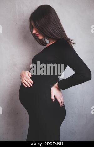 Porträt einer jungen dunkelhaarigen schwangeren Frau in einem schwarzen Kleid auf einem neutralen Hintergrund. Schwangerschaftsbump. Feminine erwartet Mutter Konzept. Stockfoto