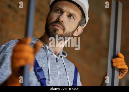 Nahaufnahme des jungen männlichen Bauherrn trägt Harthut suchen konzentriert, halten Metallbolzen für Trockenbau während der Arbeit auf der Baustelle. Haus bauen, Beruf, Sicherheitskonzept Stockfoto