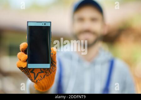 Nahaufnahme des Smartphones mit leerem Bildschirm. Junge männliche Baumeister in Uniform mit Handy, im Freien an einem warmen sonnigen Tag stehen. Technologiekonzept. Selektiver Fokus Stockfoto