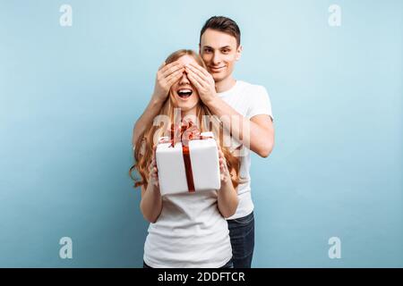 Ein gutaussehender Mann schließt die Augen seiner Frau, macht sie zu einer Überraschung und schenkt ihr vor blauem Hintergrund ein Geschenk. Valentinstag Stockfoto