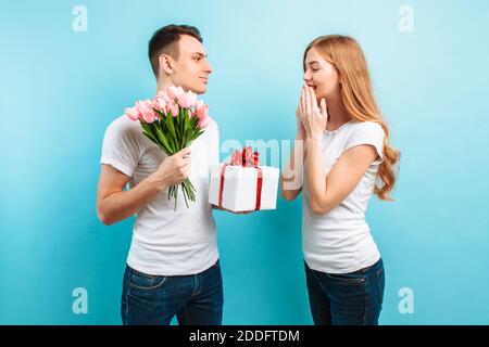 Attraktiver Mann schenkt seiner Freundin einen Blumenstrauß aus Tulpen und ein Geschenk, gekleidet in weiße T-Shirts, auf blauem Hintergrund. Valentinstag Stockfoto