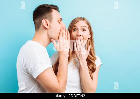 Ein Mann flüstert einer Frau ins Ohr und erzählt ihr etwas Geheimes, gute Nachrichten, auf blauem Hintergrund. Stockfoto