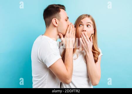 Ein Mann flüstert einer Frau ins Ohr und erzählt ihr etwas Geheimes, schockierend, auf blauem Hintergrund. Stockfoto