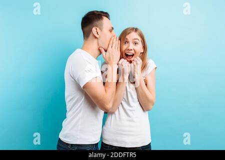 Ein Mann flüstert einer Frau ins Ohr und erzählt ihr etwas Geheimes, gute Nachrichten, auf blauem Hintergrund. Stockfoto