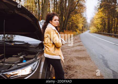 Attraktive verärgert afro behaarte Brünette neben ihrem kaputten Auto Im Wald auf leerer Straße Stockfoto