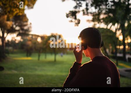 Blonde kaukasischen Jungen Musik hören mit kabellosen Kopfhörern bei Sonnenuntergang, trägt einen roten Pullover. Konzept der Technik. Musik auf der Straße. Stockfoto