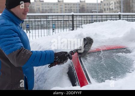 Mann in einer Winter Daunenjacke reinigt das Auto von Viel Neuschnee, der gefallen ist Stockfoto