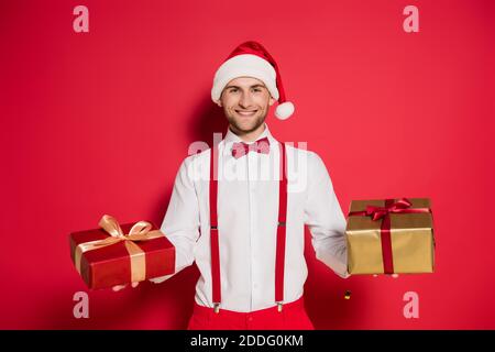 Fröhlicher Mann in santa Hut hält Geschenke auf rotem Hintergrund Stockfoto