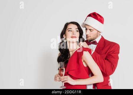 Mann in santa Hut und Fliege tragen Halskette auf Frau im Kleid mit Glas Champagner auf grauem Hintergrund Stockfoto