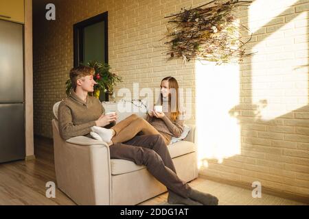 Schönes Paar in Pullover trinken Tasse Kaffee auf dem Sofa. Weihnachtsdekoration auf Hintergrund, Tannenzweige. Frau und Mann sprechen und lächeln. Stockfoto