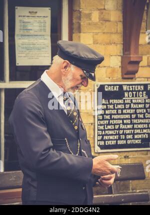 Nahaufnahme des diensthabenden Bahnhofsmeisters, der seine Taschenuhr auf dem Bahnsteig des britischen historischen Dampfbahnhofs überprüft. Stockfoto