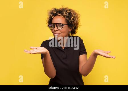 Junge afrikanische Frau mit den Armen aus, zuckerte ihre Schultern, sagte, wer kümmert sich, so was jetzt. Studio auf gelber Wand. Stockfoto