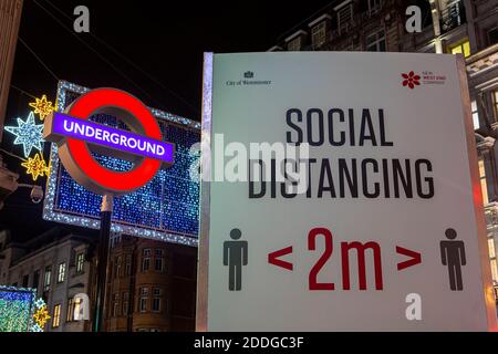 London, Großbritannien - 22. November 2020: Ein soziales Distanzierungsschild, abgebildet neben dem Londoner U-Bahn-Schild am Oxford Circus Station in London, Großbritannien. Stockfoto