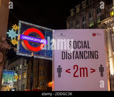 London, Großbritannien - 22. November 2020: Ein soziales Distanzierungsschild, abgebildet neben dem Londoner U-Bahn-Schild am Oxford Circus Station in London, Großbritannien. Stockfoto