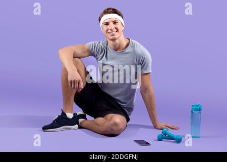 In voller Länge Porträt des jungen athletischen Mann mit Hanteln und Eine Flasche Wasser sitzt auf einem fliederfarbenen Studiohintergrund Stockfoto