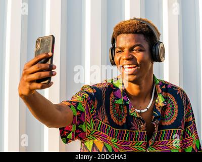 Junge positive afroamerikanische Männer in Kopfhörer und trendige Kleidung Auf der Straße stehen und Selfie auf dem Smartphone machen Stockfoto