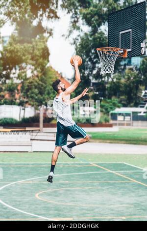 Ganzkörper-Seitenansicht des entschlossenen jungen männlichen Basketballspielers In der Sportbekleidung springen und schießen Ball in der Nähe Reifen während des Trainings Allein auf der Straße Playgr Stockfoto