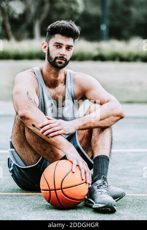Ganzer Körper von schweren muskulösen jungen bärtigen Basketballspieler in Sportbekleidung, die neben dem Ball auf dem Spielplatz auf der Straße sitzt und anschaut Kamera Stockfoto