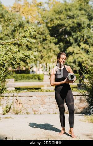 Ganzkörper aus positiv schlank barfuß weiblich in schwarzer Sportbekleidung Halten gerollte Matte während der Vorbereitung für Yoga-Praxis in sonnigen Sommerpark Stockfoto