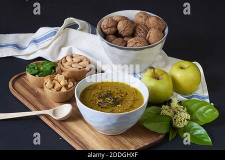 Von oben leckere vegane Cremesuppe mit Kürbiskernen Auf Holztablett mit Brotkruten und frischem Basilikum angeordnet Platziert auf dem Tisch neben Äpfeln und wa Stockfoto