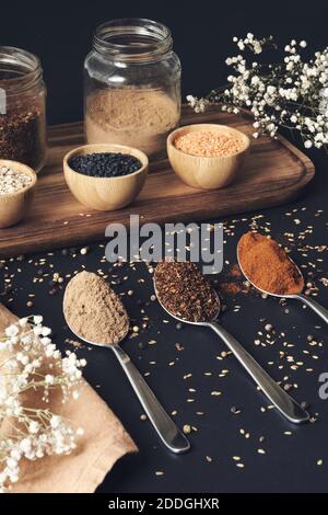Draufsicht auf verschiedene Körner in Gläsern und aromatischen Gewürzen In Löffeln auf Holztablett auf schwarzem Tisch angeordnet Stockfoto